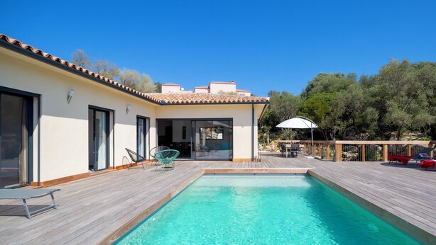 Una piscina con cubierta de madera, un área de estar al aire libre techada y un edificio con ventanas y puertas grandes.