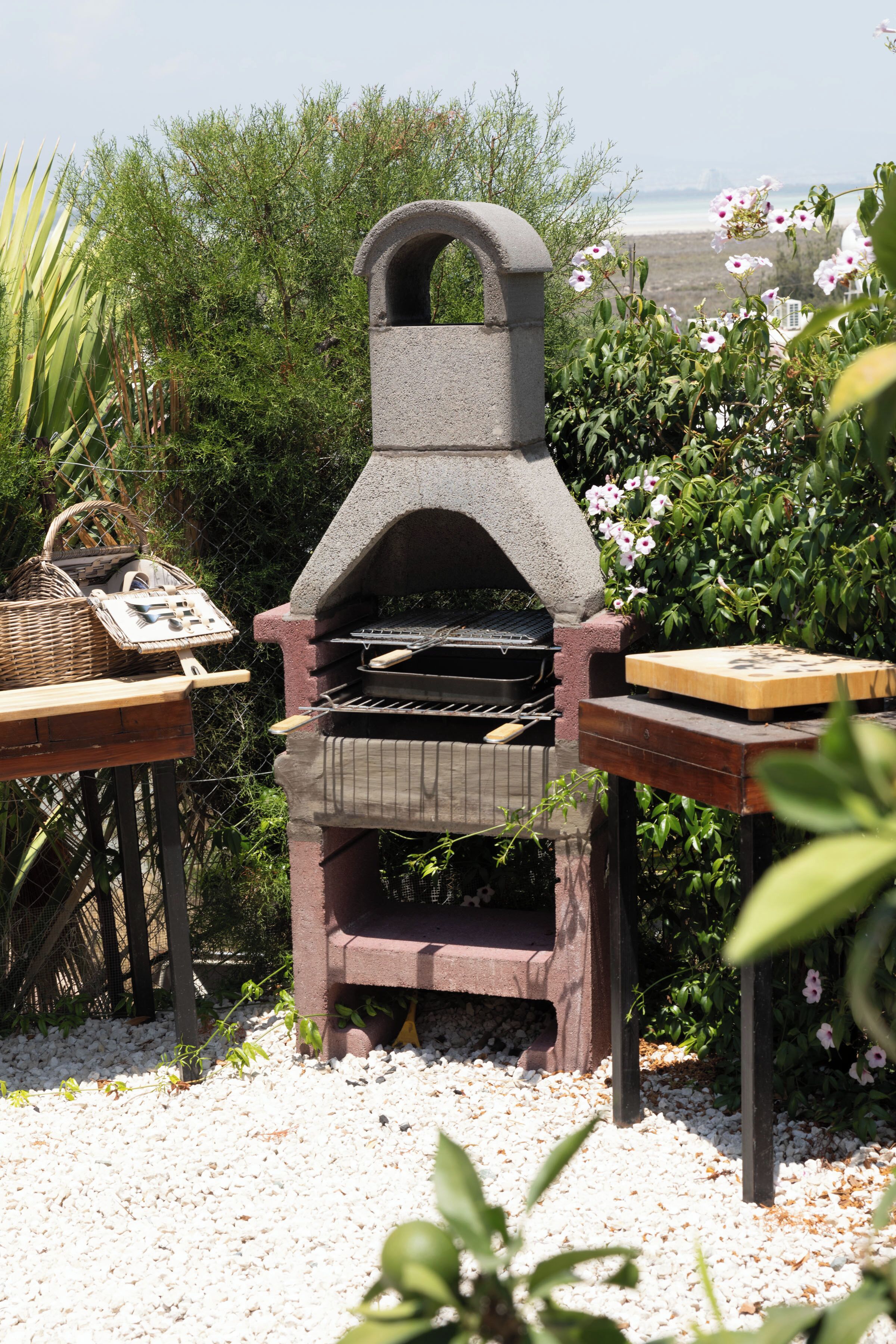 Un barbecue extérieur en pierre, doté d’une cheminée, entouré de verdure et de petits galets blancs.