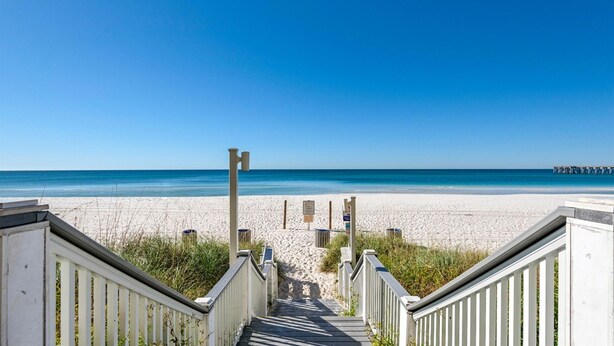 En trätrappa leder ner till en sandstrand med en klarblå himmel och en brygga i fjärran.