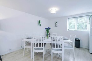 A white dining area with a table and chairs, a small plant, and a window with blinds.