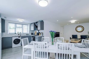 A modern kitchen with a built-in washing machine, a dining area with white furniture, and a living space with a sofa and a round mirror. - Stunning 3BR for Summer Leisure | Shrewsbury Town (Shropshire)