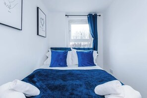 Ein Schlafzimmer mit einem Bett, zwei Kissen, einem Fenster mit Vorhängen und zwei gerahmten Bildern an der Wand.