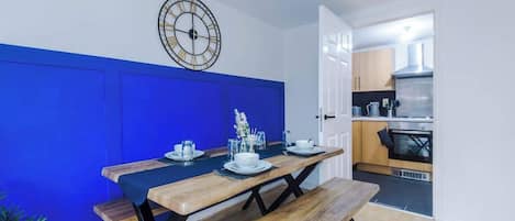 Un coin repas avec une table en bois dressée pour deux personnes, un mur d’accent bleu et une cuisine visible à travers une porte ouverte.