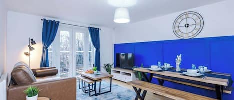 A modern living room with a sofa, a dining table with benches, a flat-screen TV, and a blue accent wall.