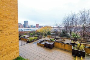 Eine Dachterrasse mit Blick auf eine Stadtlandschaft, mit einer Ziegelwand und einem gepflasterten Bereich mit Pflanzgefäßen.
