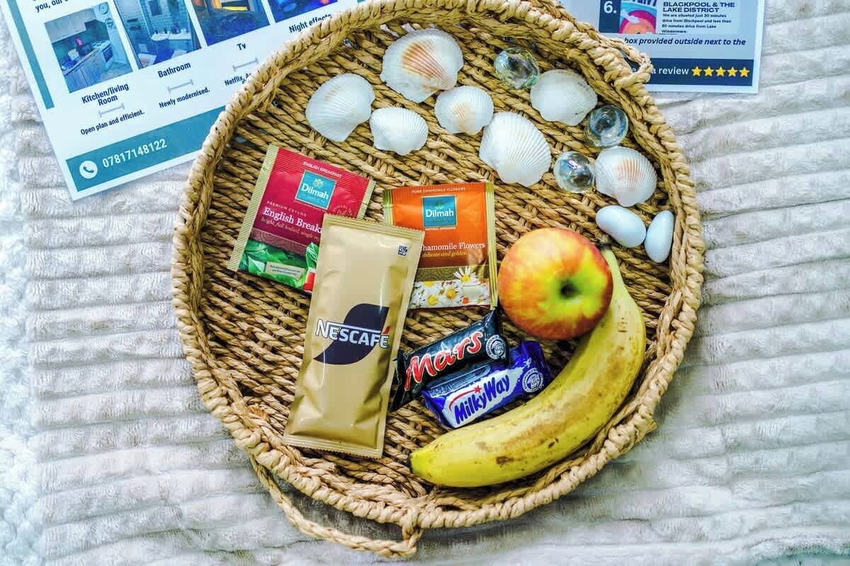 A wicker basket containing a banana, a Mars chocolate bar, a Nescafe packet, a packet of English Breakfast tea, a packet of chamomile flowers, and several seashells.