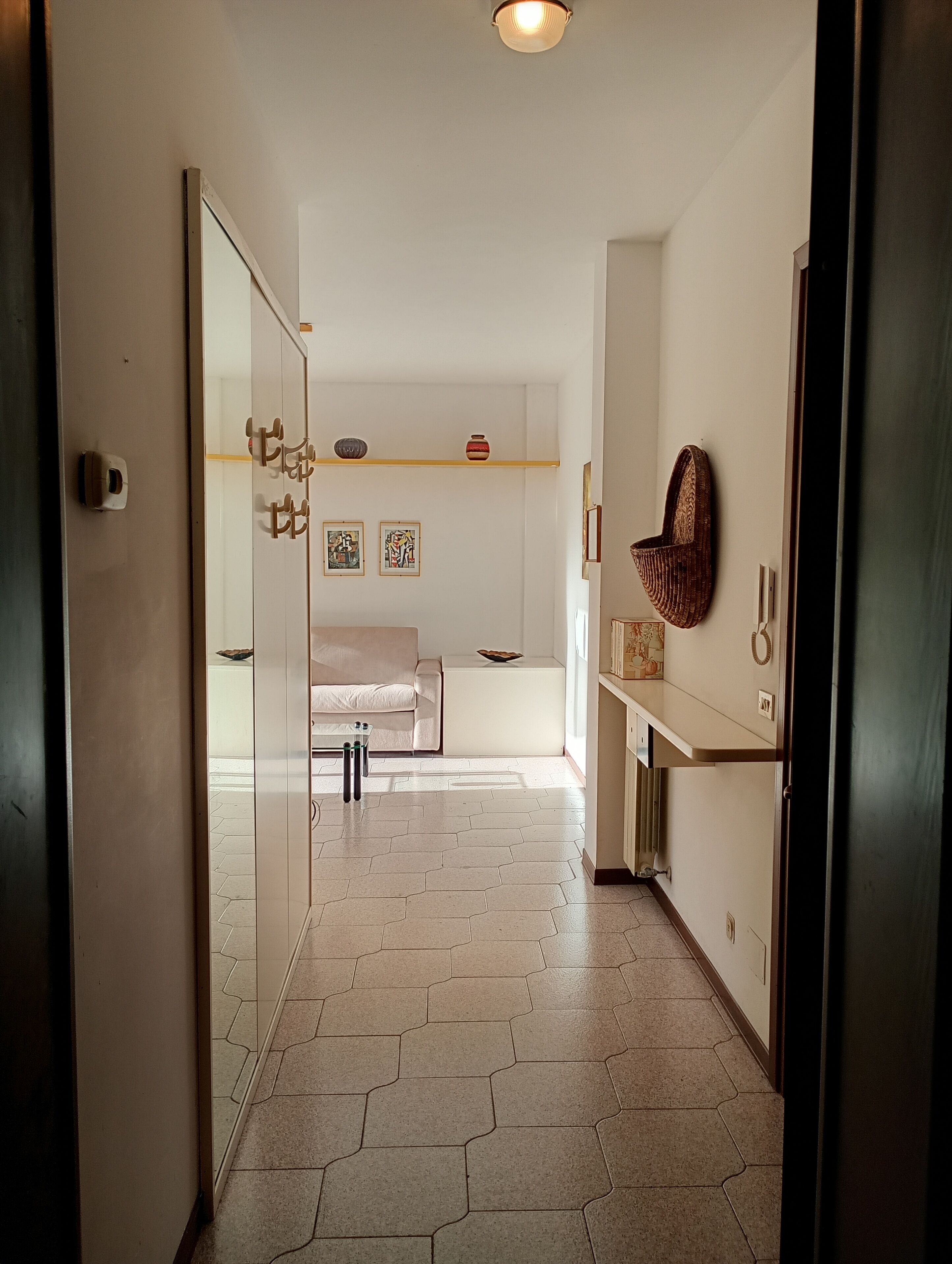 Un corridoio con una porta in vetro, un divano bianco, un tavolino e una mensola con oggetti decorativi.
