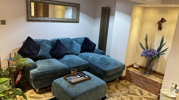 A living room with a sectional sofa, a small ottoman, a mirror, and a decorative plant. - Wonky 2 A Cosy Hideaway in the Heart of Cheshire (Macclesfield)