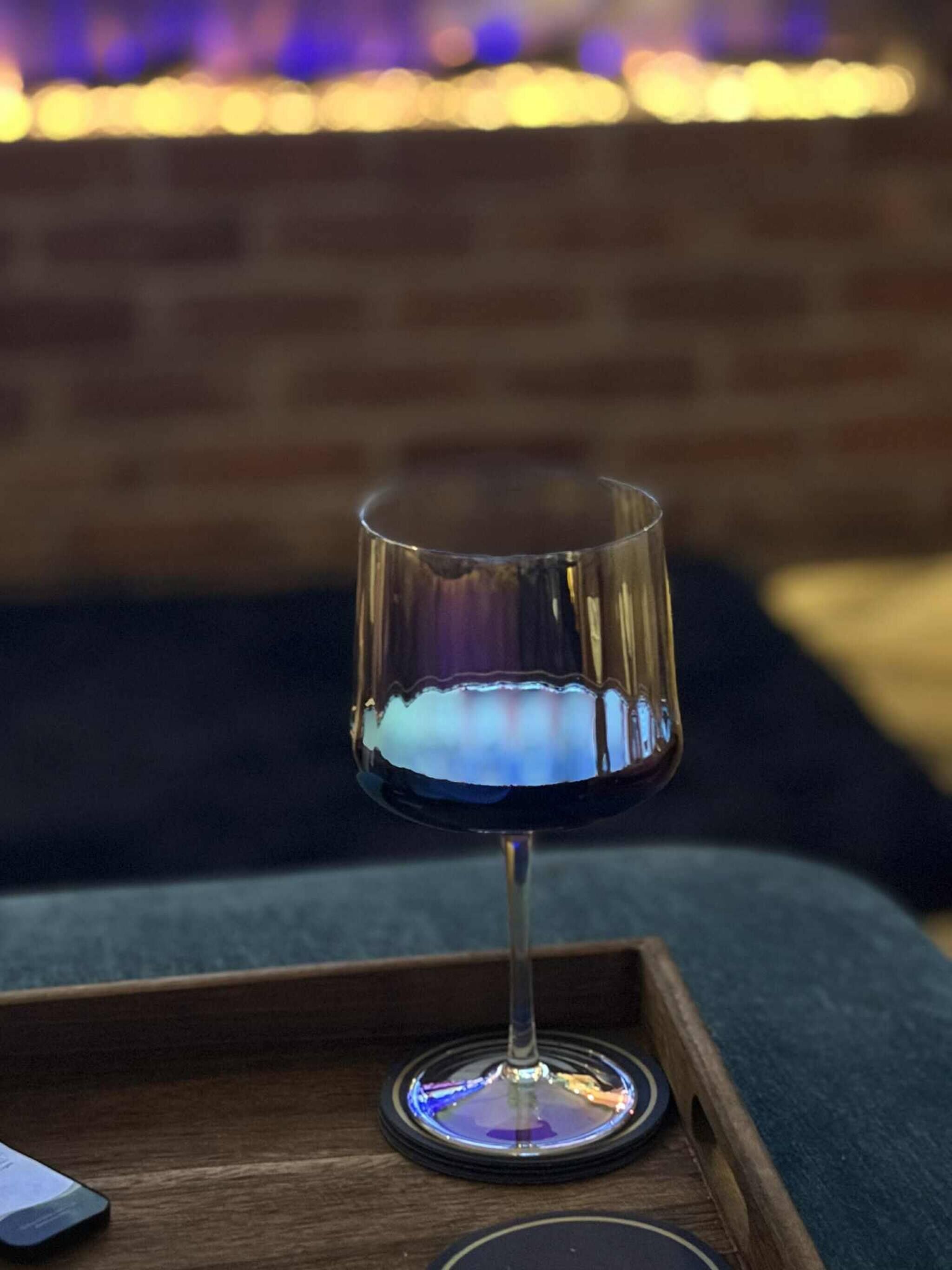 A glass of red wine on a wooden tray with a coaster, placed on a couch with a brick wall and string lights in the background.