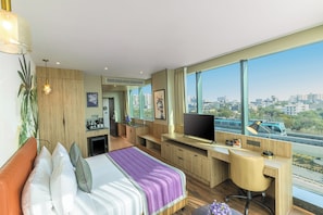 A modern hotel room with a bed, desk, TV, and large windows offering a city view.