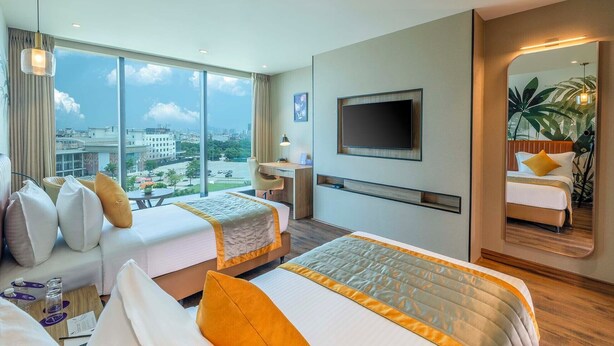 Habitación de hotel con dos camas, televisión, un ventanal grande y vistas a la ciudad, y un espejo.