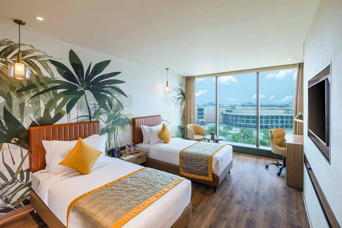 A hotel room with two beds, a large window with a city view, a desk with a chair, and a large plant.
