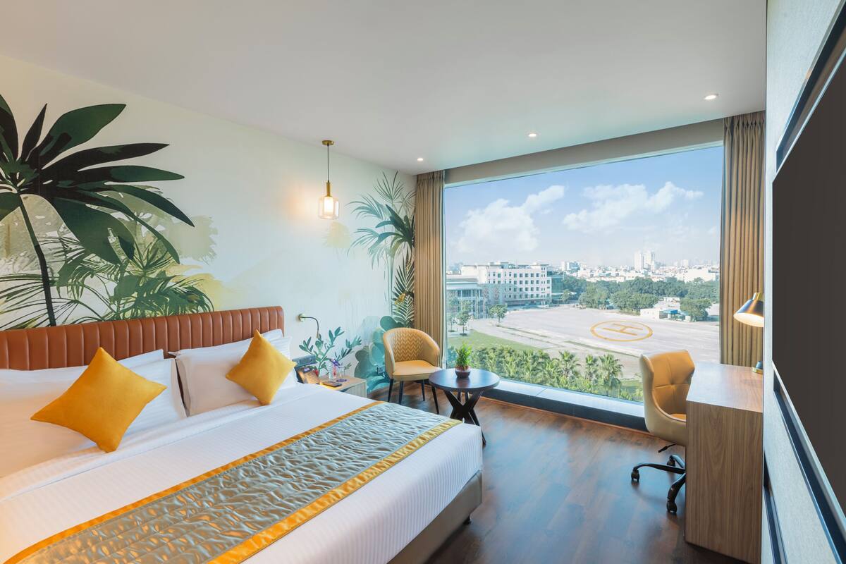 A hotel room with a large bed, a desk, a chair, a view of the city, and a large plant.