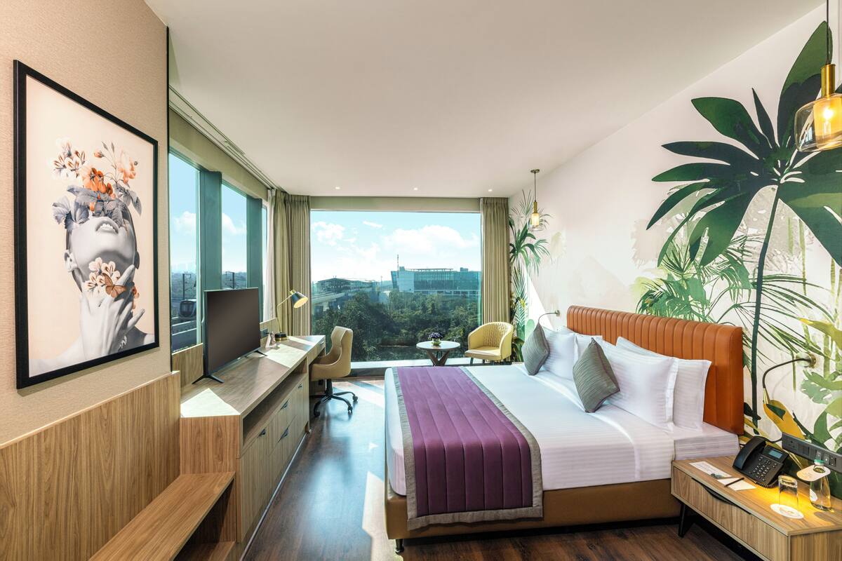 A hotel room with a bed, desk, chair, TV, and a large window with a city view.