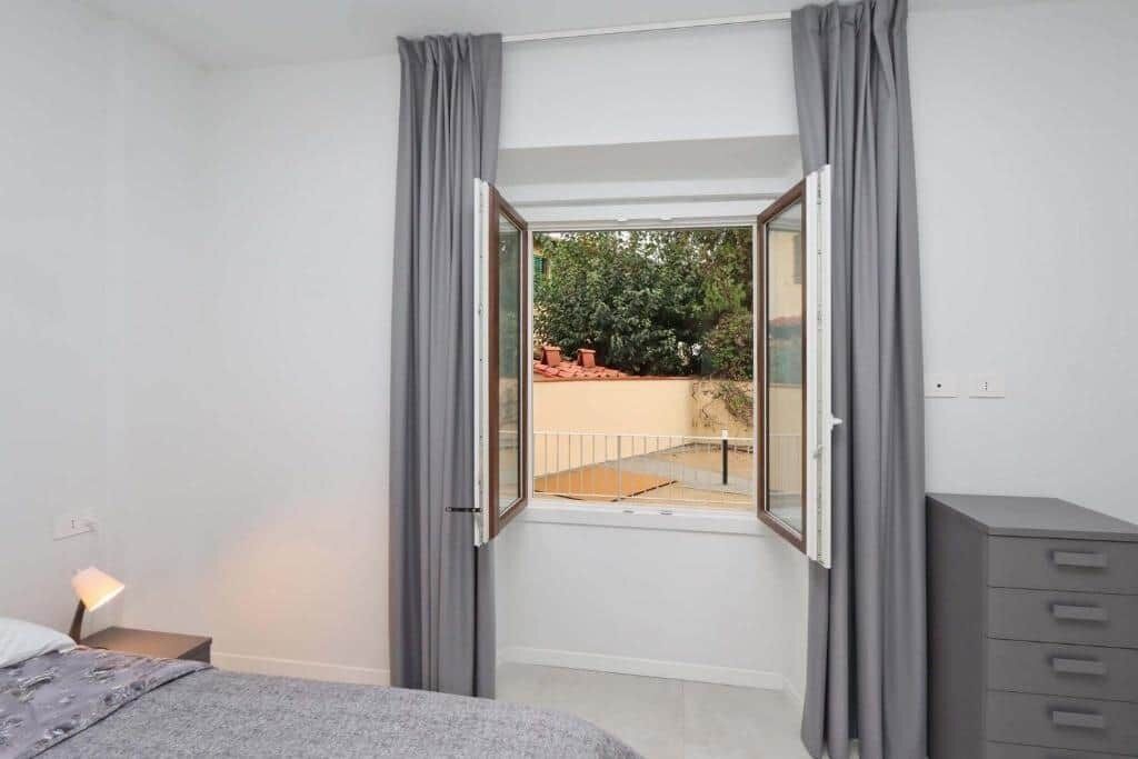 Ein Schlafzimmer mit großem Fenster, einem Bett, einem Nachttisch und einer grauen Kommode.