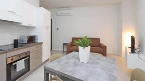 Cozinha compacta com eletrodomésticos modernos, área de jantar com mesa e cadeiras, e sala de estar com sofá e planta.