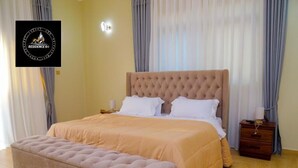 A bedroom with a large bed, a matching bench, a nightstand, and a wall-mounted lamp. - Residence Six Sigma (Entebbe)