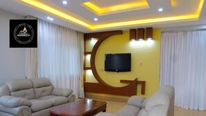 A living room with two beige sofas, a wooden coffee table, a flat-screen TV mounted on the wall, and a decorative wall piece. - Residence Six Sigma (Entebbe)