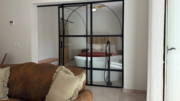 Una sala de estar moderna con un sofá marrón y un cojín a rayas, una puerta corrediza de vidrio que da a un dormitorio con una cama y una bañera blanca.