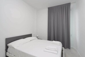 Ein Einzelbett mit weißer Bettwäsche, einem grauen Kopfteil und einem grauen Vorhang an einem Fenster.