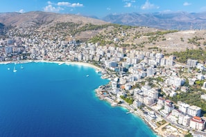 An aerial view of a coastal city with dense urban development, a marina, and a mountainous backdrop. - Panorama View Apartment In Saranda - 271 (Sarandë)
