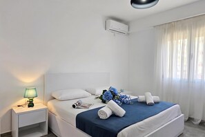 A hotel room with a bed, a nightstand, a lamp, a magazine, a bouquet of flowers, and a window with curtains. - Holiday Apartment In Saranda - 003 With Balcony (Sarandë)