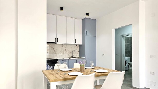 A modern kitchen with white cabinets, a marble backsplash, and a dining area with a wooden table and white chairs. - Holiday Apartment In Saranda - 003 With Balcony (Sarandë)