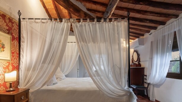 Una cama con dosel adornada con cortinas blancas, una mesita de noche de madera con una lámpara y un cuadro enmarcado en la pared.