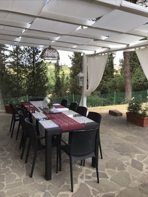 An outdoor patio with a covered table and chairs, surrounded by stone flooring and greenery.