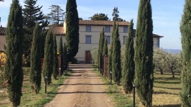 Un chemin de terre mène à un bâtiment dont le toit est recouvert de tuiles, et qui est bordé de grands cyprès.