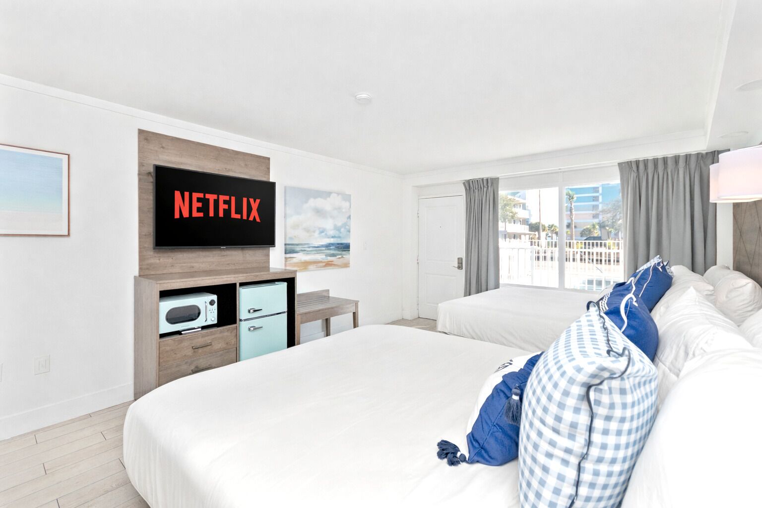 Ein Hotelzimmer mit einem großen Bett, einem Einzelbett, einem an der Wand befestigten Fernseher, einem Schreibtisch mit Computer und einem Fenster mit Blick auf Gebäude.
