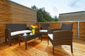 Eine Holzterrasse mit Korbmöbeln, darunter eine Couch und Stühle, sowie ein kleiner Tisch mit einer gelben Vase und einer weißen Tasse.