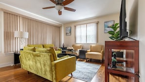 A living room with a yellow sofa, a ceiling fan, a TV on a wooden stand, and a window with blinds. - 5 Woods Dr (Mechanicsburg)