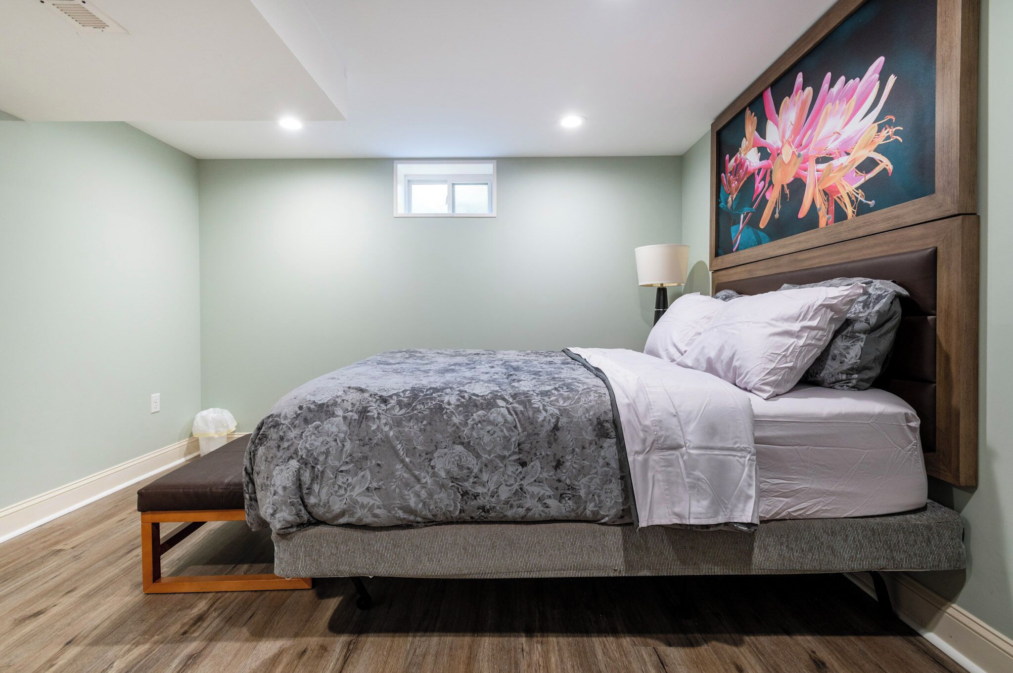 Un dormitorio con cama, una banca, mesita de noche, lámpara y ventana.