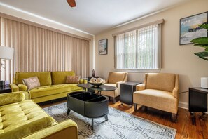 Una sala de estar con un sofá amarillo, un sillón beige, una mesa de centro negra y un ventanal con persianas.