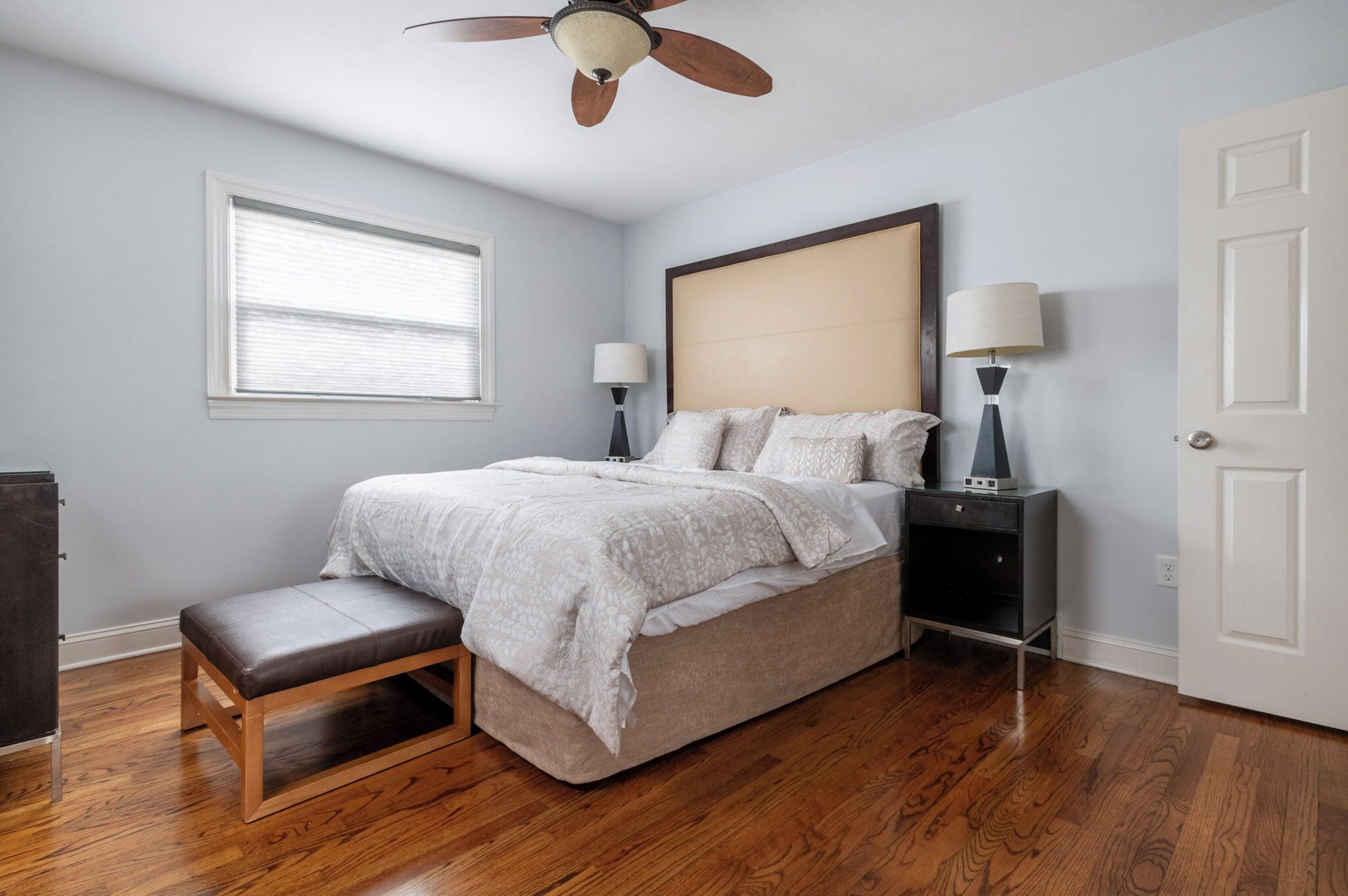 A bedroom with a bed, bedside tables, a bench, a ceiling fan, a window, and a door.