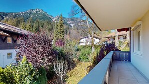 Uma varanda com vista para as montanhas, uma casa com telhado inclinado e um jardim com diversas plantas.