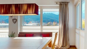 Uma sala de jantar com mesa e cadeiras de madeira, um banco com almofadas vermelhas, uma grande janela com vista para montanhas e uma cortina à direita.
