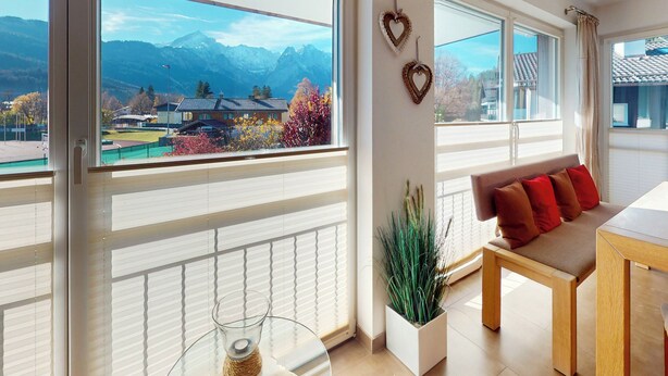 Quarto moderno com grandes janelas, vista para montanhas, sofá com almofadas e um vaso de plantas.