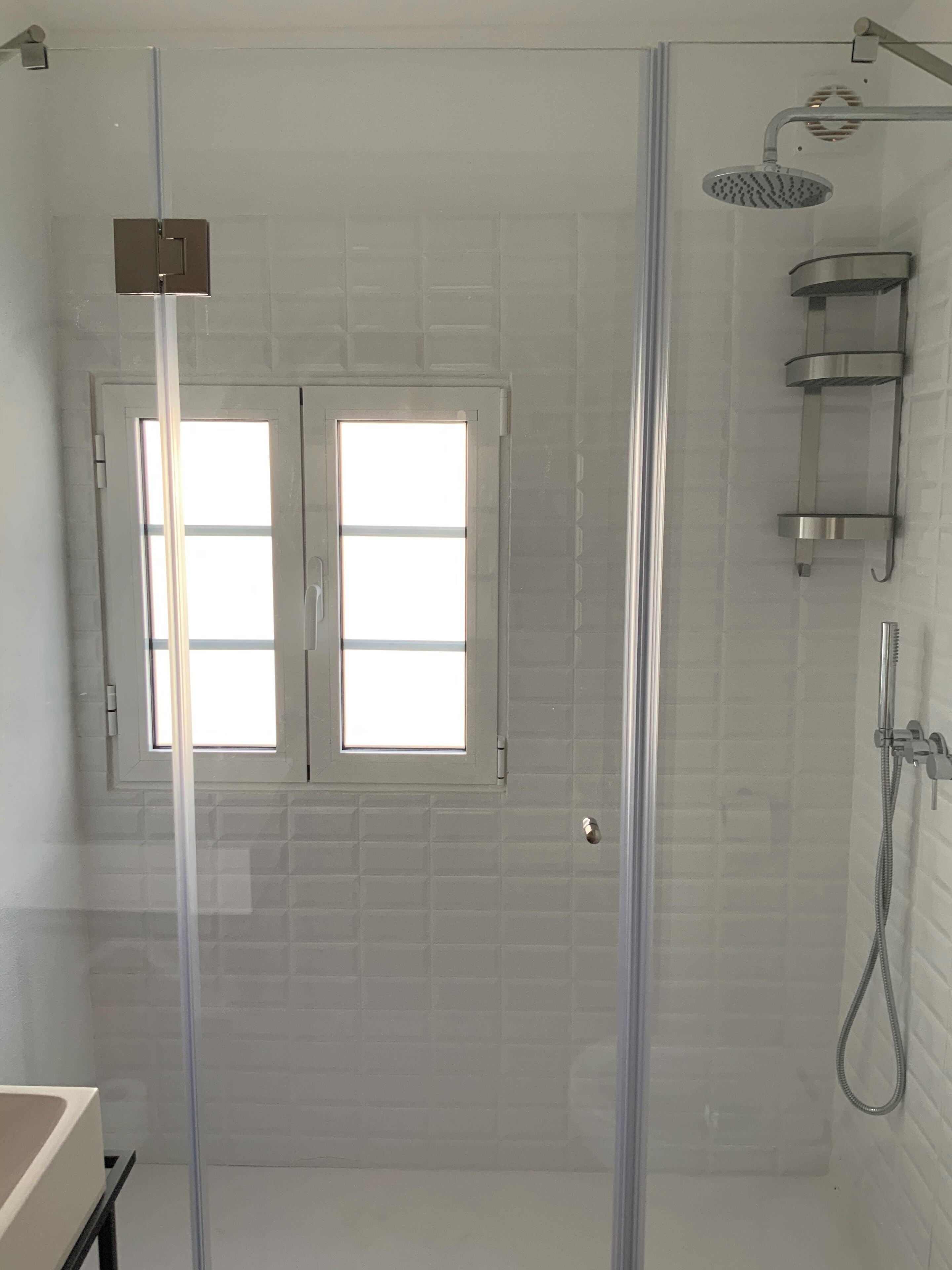 A modern bathroom with a glass-enclosed shower, a white bathtub, and a window with blinds.
