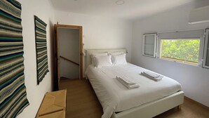 A bedroom with a bed, a window with a view of greenery, a door, and a wall with a patterned curtain. - Casa Carmo (Évora)