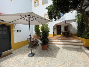 Une cour avec une table et des chaises placées sous un grand parasol, un pot de fleurs et un escalier menant à une entrée.