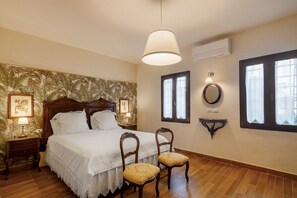 Quarto com cama, duas cadeiras, mesa de cabeceira, luminária na parede e janela com cortinas.