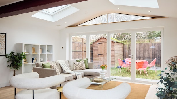 Ein modernes Wohnzimmer mit einem Ecksofa, weißen Sessel und einem Glastisch. Ein Bücherregal ist mit Dekoration und einer Pflanze dekoriert. Große Fenster bieten Blick auf eine Holzterrasse mit Gartenmöbeln.