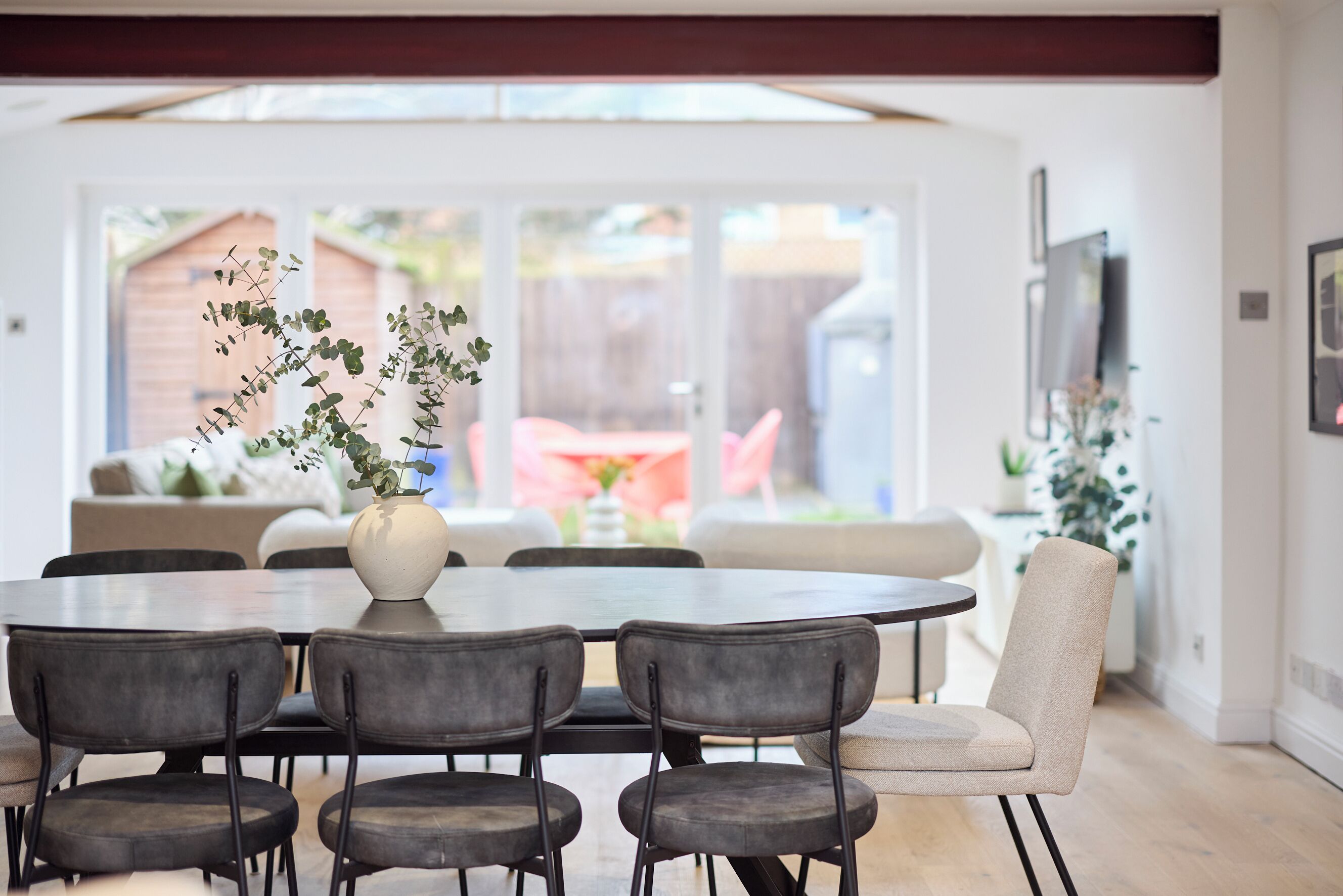 A dining area with a round table and six chairs, a vase with greenery, and a view of a backyard with a wooden fence and outdoor seating.