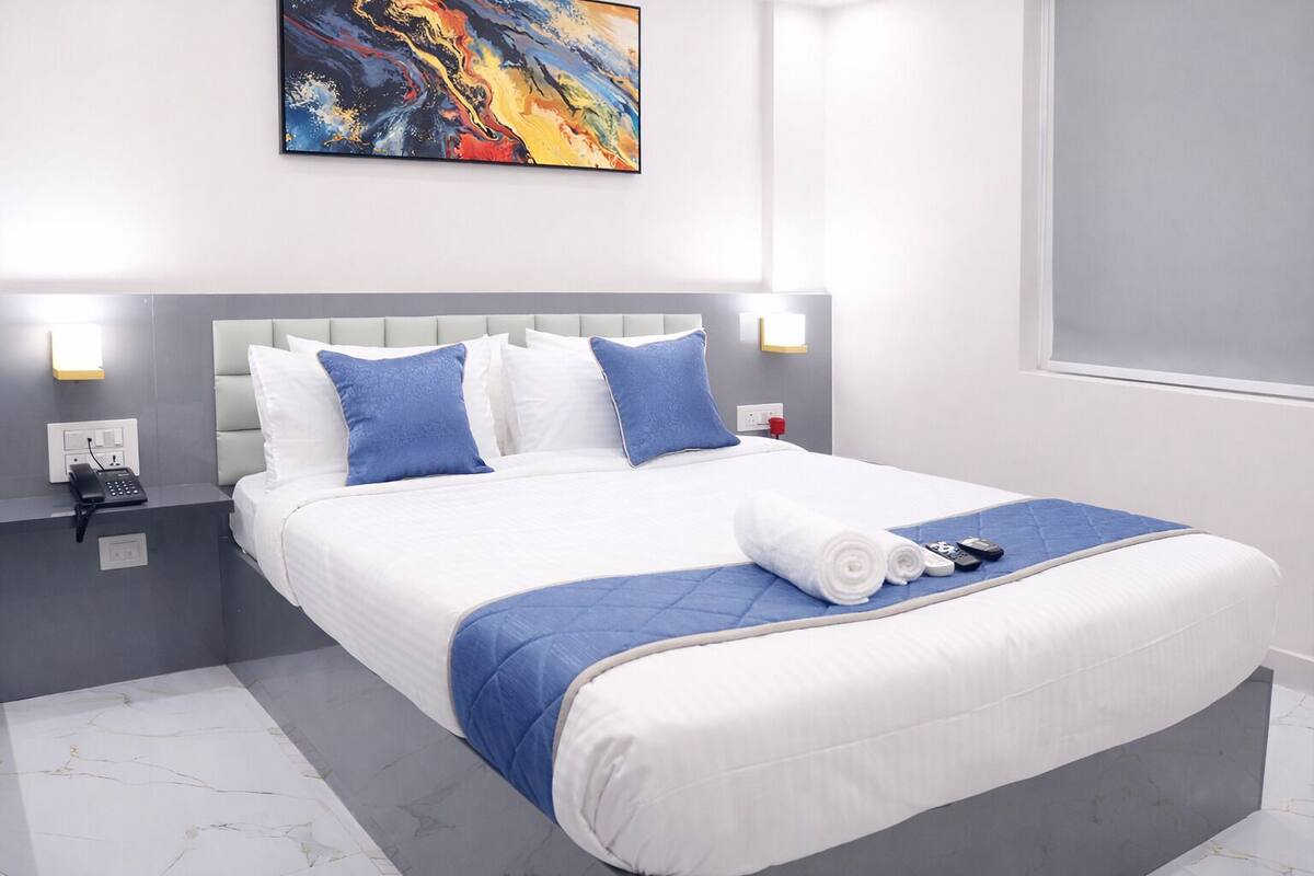 A modern hotel room with a large bed, blue bedding, a marble floor, a painting on the wall, and a bedside table with a phone and a red cup.