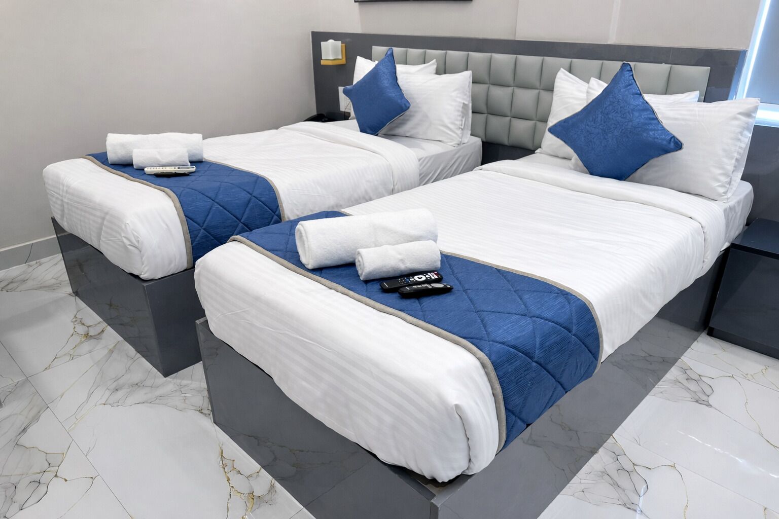 A hotel room with two beds, each with white bedding and blue accents, a grey headboard, and marble flooring.