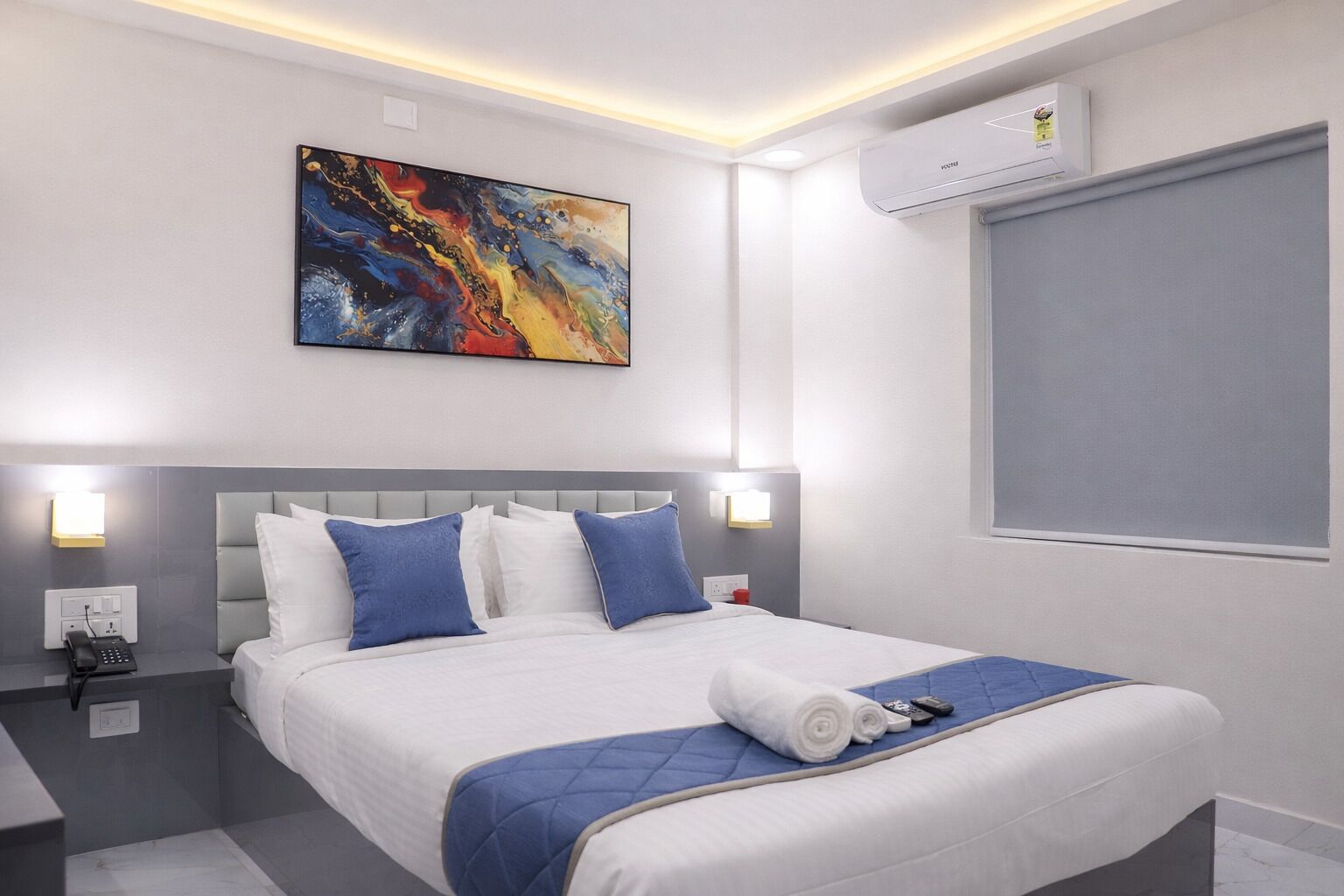 A modern hotel room with a large bed, a painting above the bed, bedside lamps, an air conditioning unit, and a window.