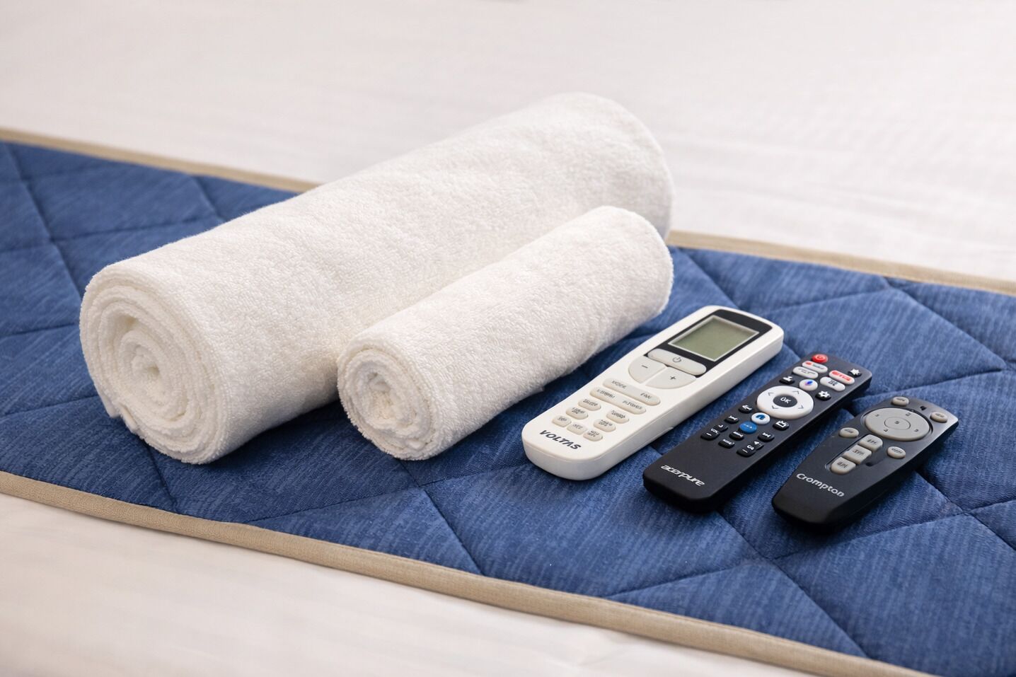 Un lit recouvert d’une courtepointe bleue, deux serviettes blanches pliées et deux télécommandes.