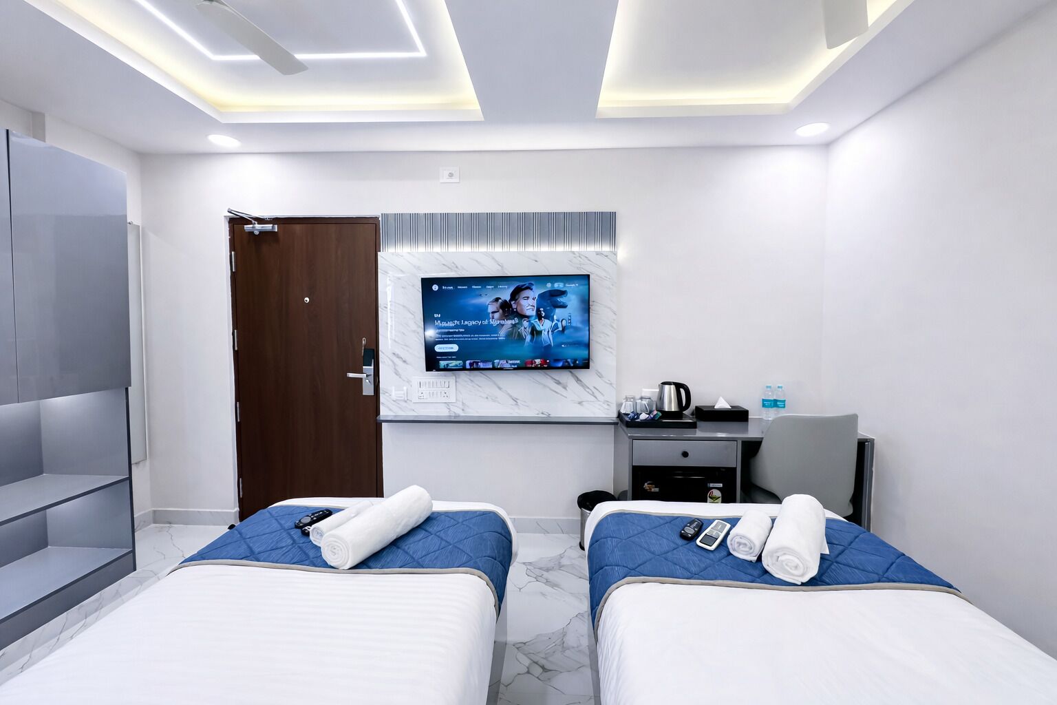 A hotel room with two beds, a TV mounted on the wall, a desk with a chair, and a wardrobe.